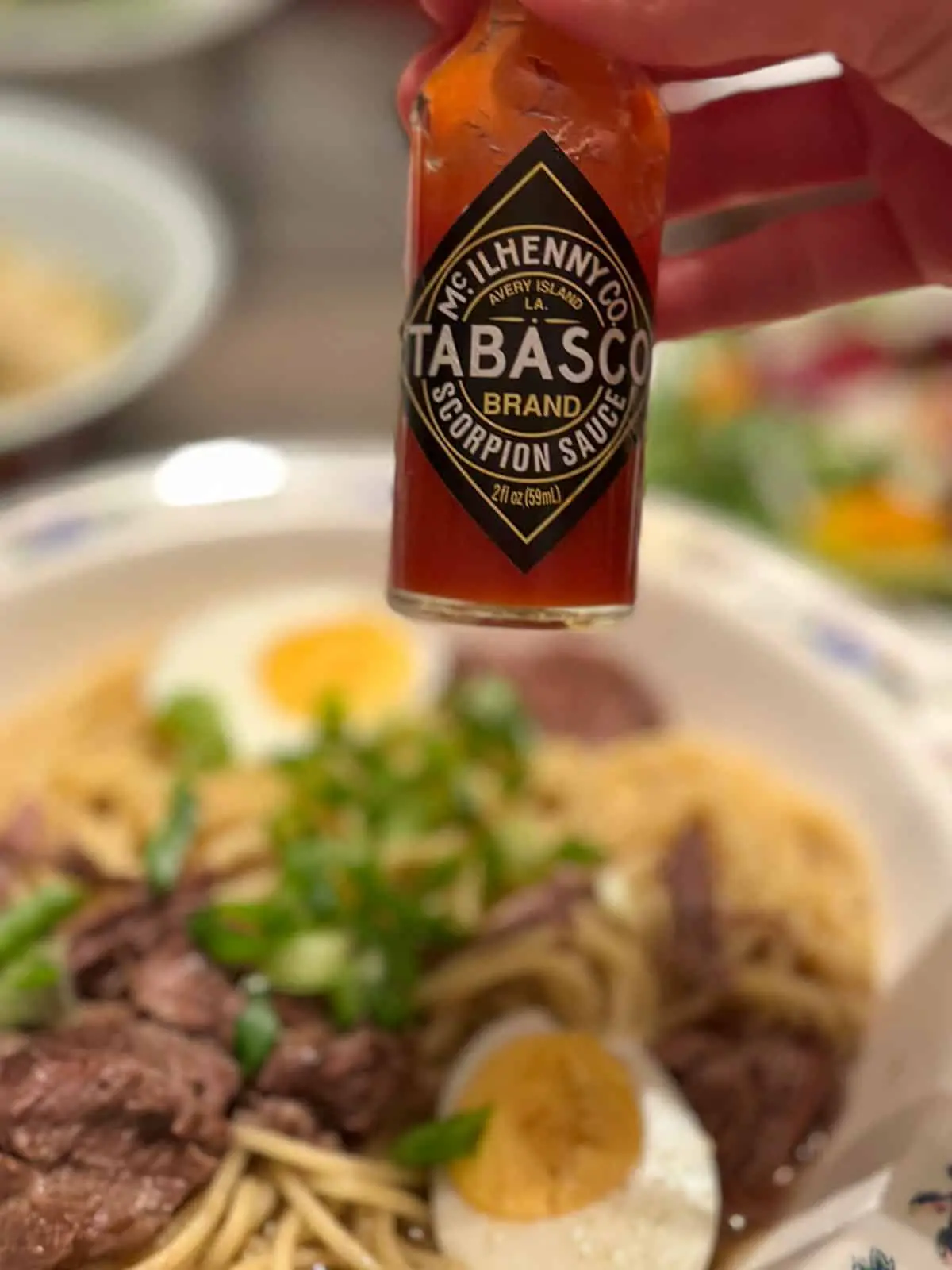 A bowl containing yakamein which is shredded beef and noodle soup garnished with hard boiled eggs and green onions and someone holding a bottle of Tabasco Scorpion Sauce over the bowl.