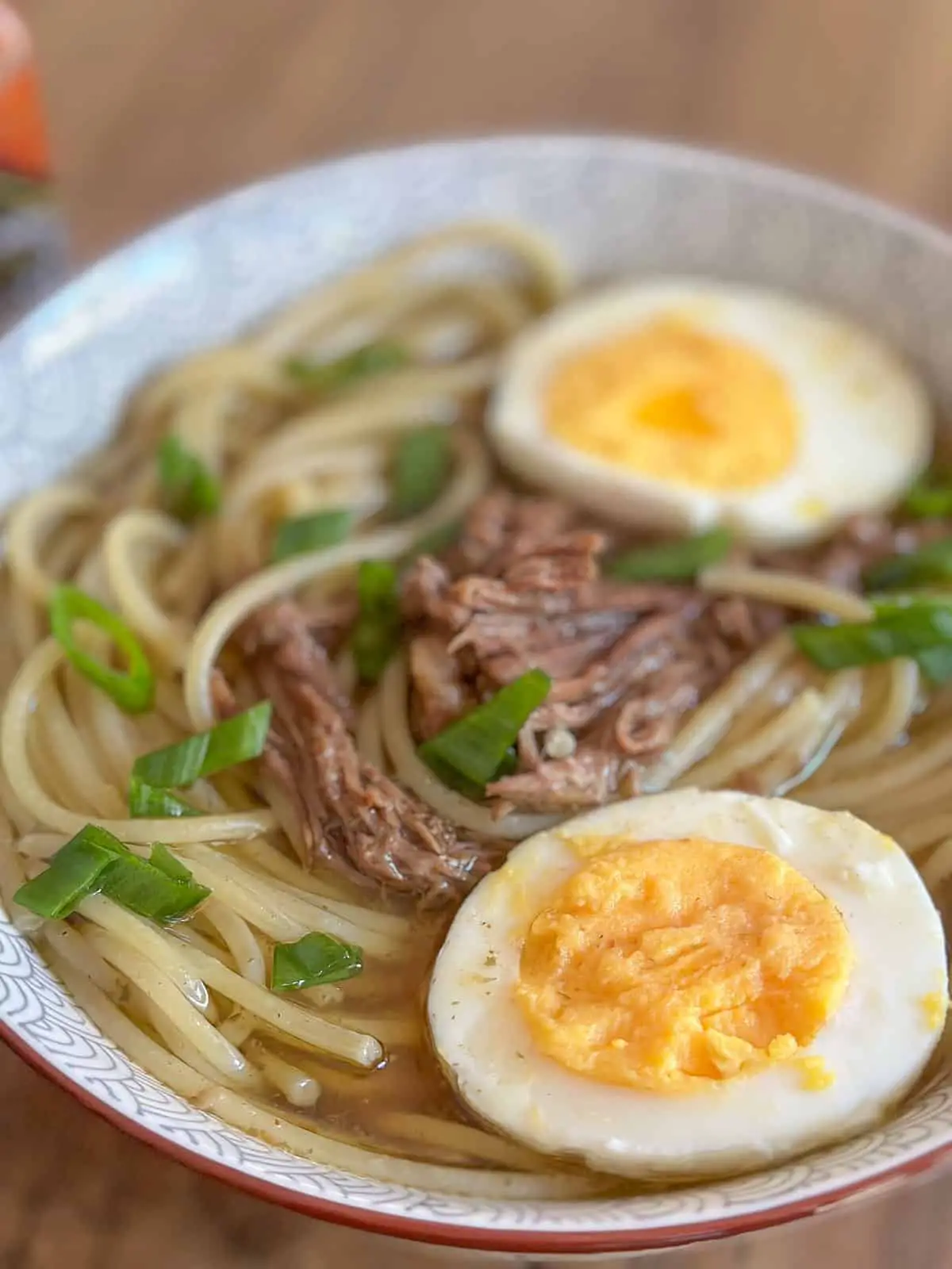 Shredded beef and noodles in a beef broth with hard boiled eggs which have been halved and green onions garnishing the noodle soup which was placed in a soup bowl with a bottle of Tabasco in the background.