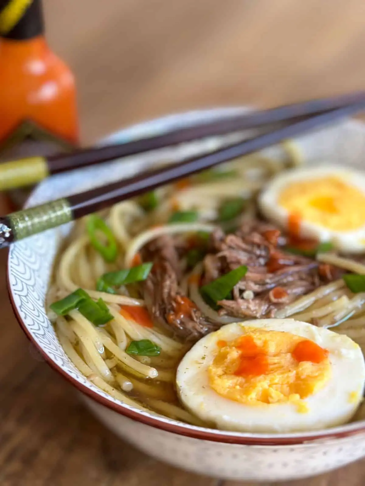 Shredded beef and noodles in a beef broth with hard boiled eggs which have been halved and green onions garnishing the noodle soup which was placed in a soup bowl with chopsticks resting on the bowl with a bottle of Tabasco in the background and splashes of Tabasco on the soup.