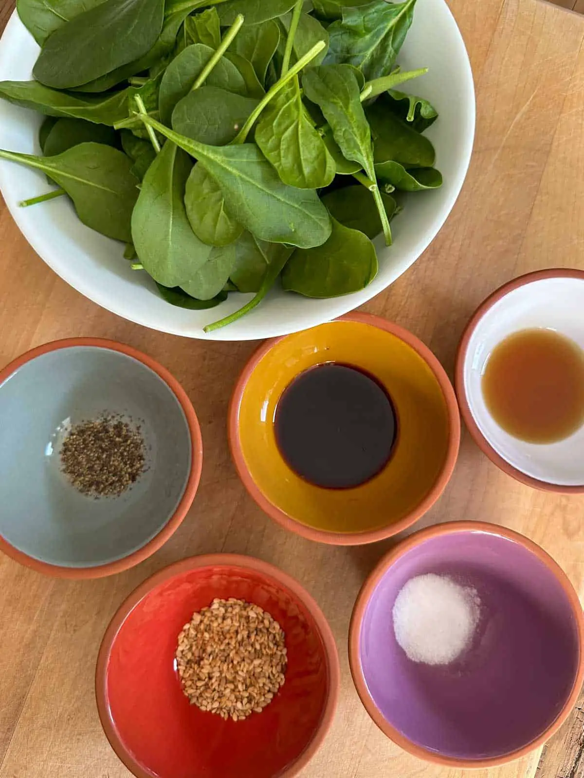Raw baby spinach in a white bowl, small colored bowls filled with pepper, soy sauce, sesame oil, sesame seeds, and sugar respectively.