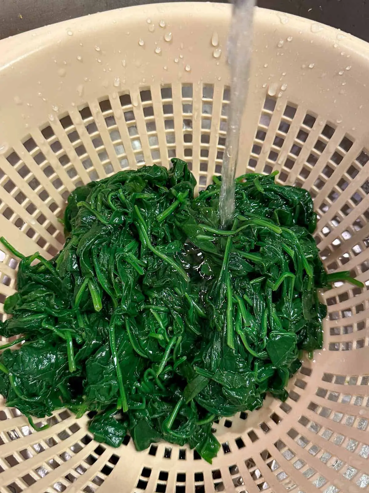 Wilted spinach in a colander with a stream of water running down onto the spinach.