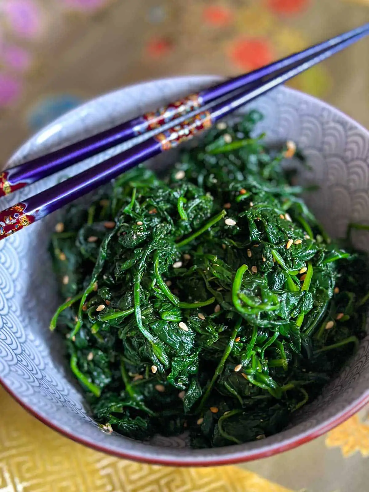 Asian style spinach flecked with sesame seeds placed in a patterned bowl with a pair of purple chopsticks resting on the bowl.