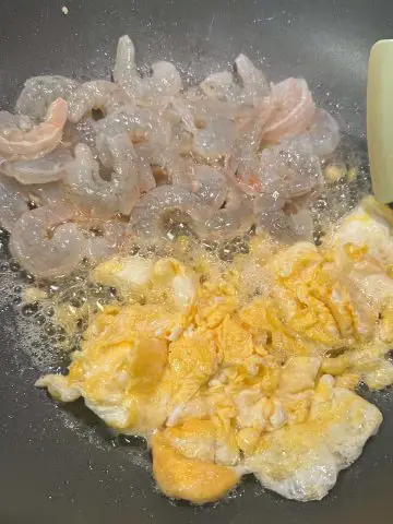 Egg and shrimp in oil cooking in a wok with a hint of a blue utensil on the top right side.