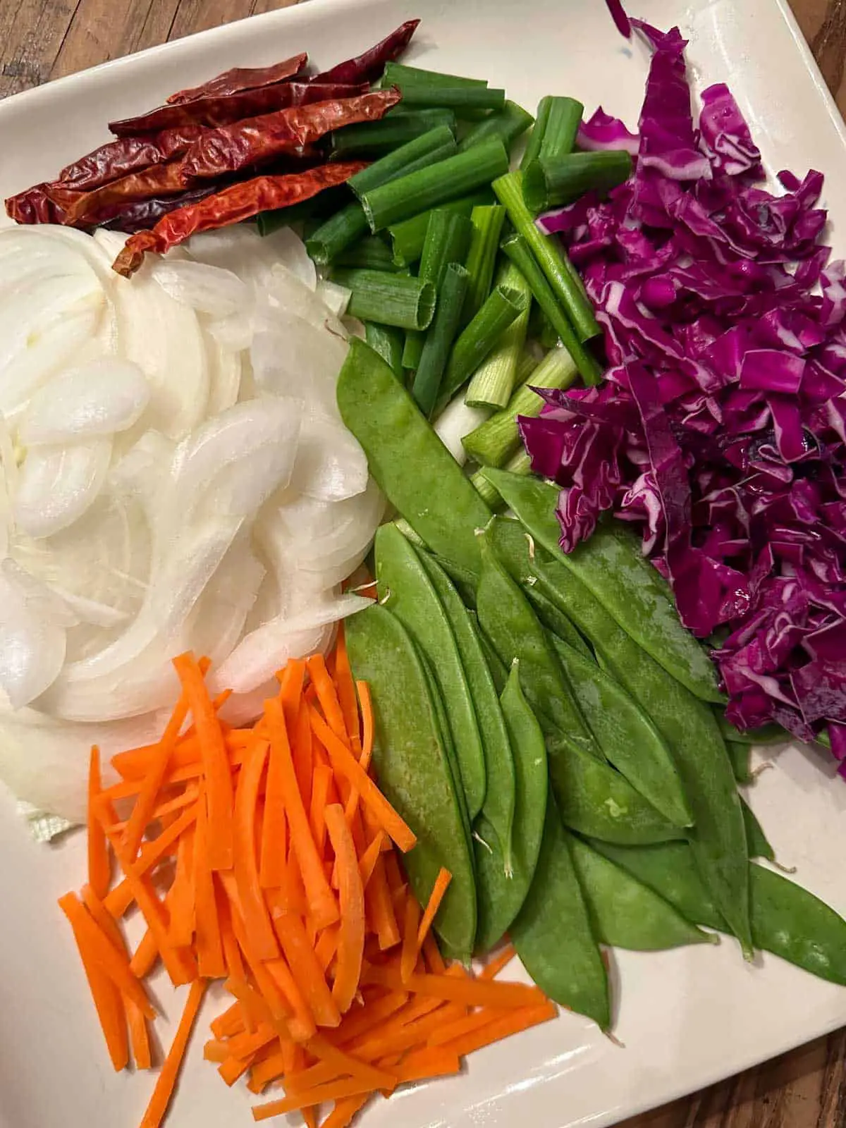Sliced onion, julienned carrots, snow peas, sliced cabbage, sliced green onions and Asian dried chilies on a white plate.