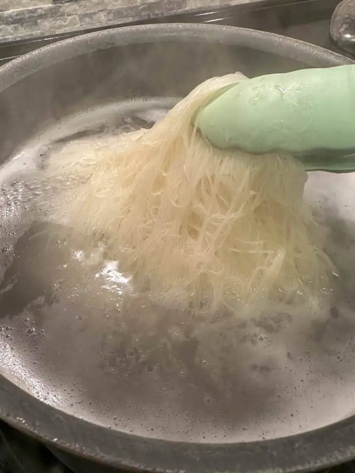 Rice vermicelli noodles cooking in water in a pan with a pair of blue tongs holding some of the noodles out of the water.