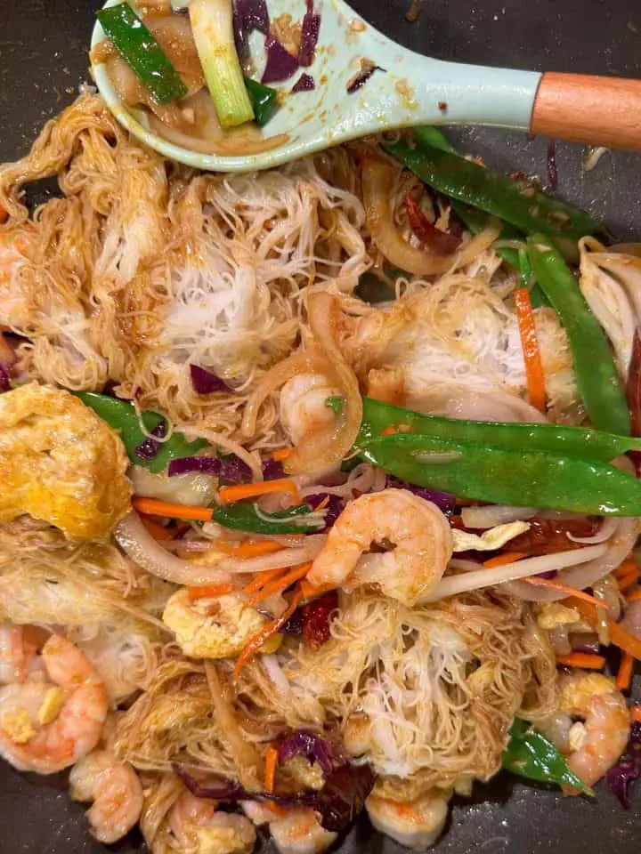 Rice noodles, shrimp, snow peas, green onions, cabbage, carrots, egg, and dried chilies in a wok with a blue silicone spoon with wooden handle resting on the side of the wok. There are some green onions, onion, and cabbage in the spoon.
