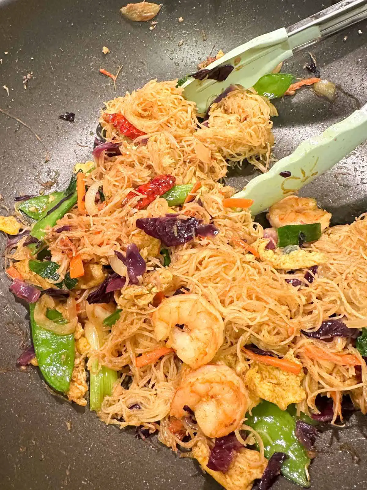 Rice noodles, shrimp, snow peas, cabbage, carrots, egg, and dried chilies in a wok with a pair of blue silicone tongs resting on the side of the wok.