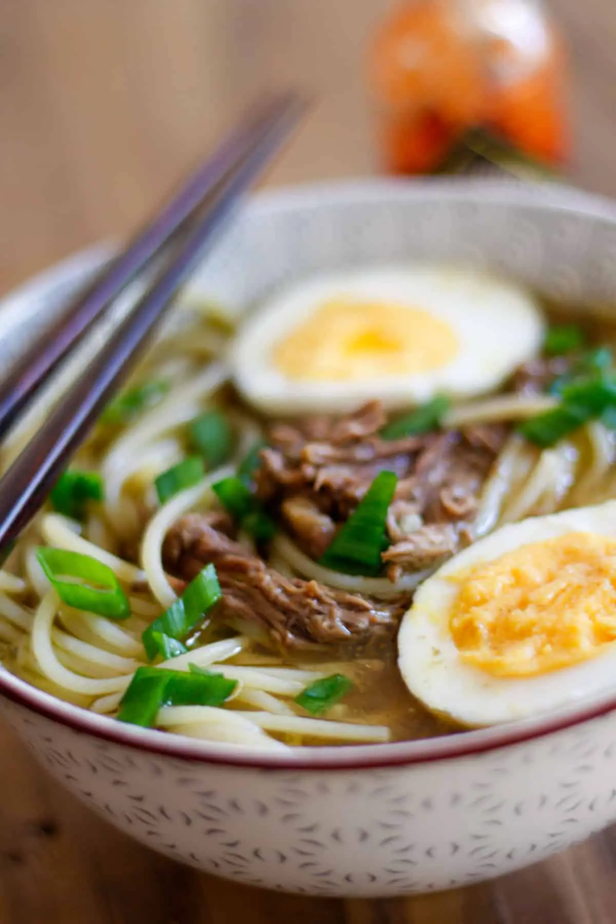 Shredded beef and noodles in a beef broth with hard boiled eggs which have been halved and green onions garnishing the noodle soup which was placed in a soup bowl with chopsticks resting on the bowl and a bottle of Tabasco in the background.
