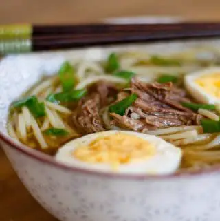 Shredded beef and noodles in a beef broth with hard boiled eggs which have been halved and green onions garnishing the noodle soup which was placed in a soup bowl with chopsticks resting on the bowl.