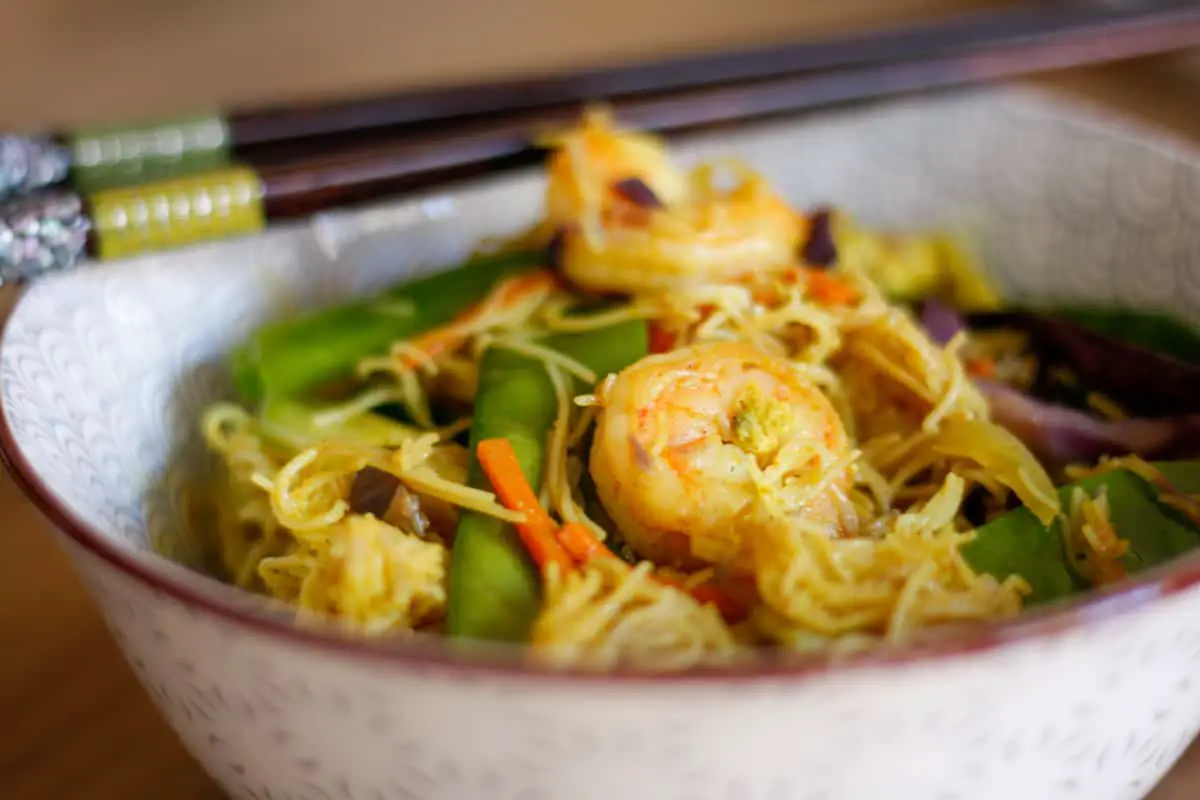 Shrimp Mei Fun with ingredients rice vermicelli noodles with snow peas, shrimp, Asian chilies, carrots and onion in a bowl with chopsticks resting on the side of the bowl.