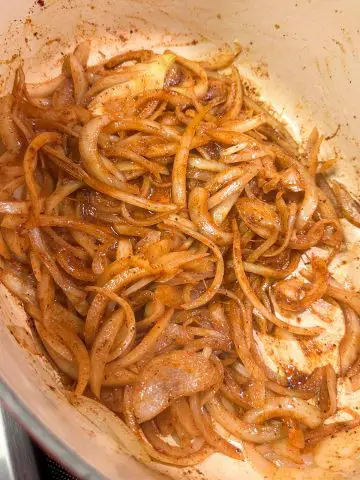 Sliced onions in a dutch oven tossed with seasonings so that they are a reddish color.