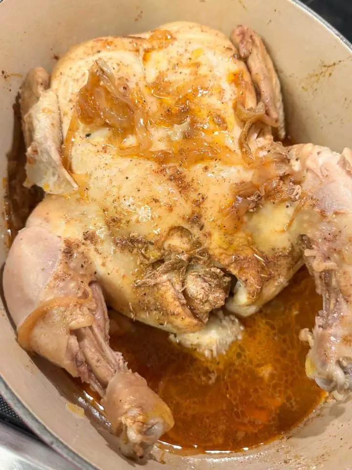 A whole chicken at the bottom of a Dutch oven. There is brown colored braising liquid and sliced onions in the dutch oven as well.