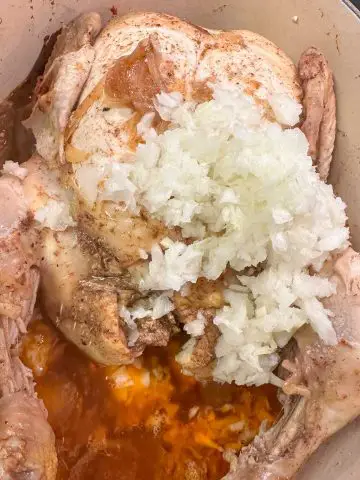 A whole chicken at the bottom of a dutch oven topped with lots of diced onion. There is an orange colored liquid which is fat in the dutch oven.