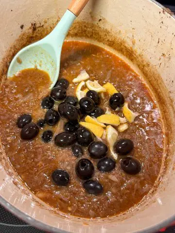 A dutch oven containing a dark sauce rich with onions, olives, and pieces of lemon rind. There is a blue spoon with a wooden handle resting in the dutch oven.