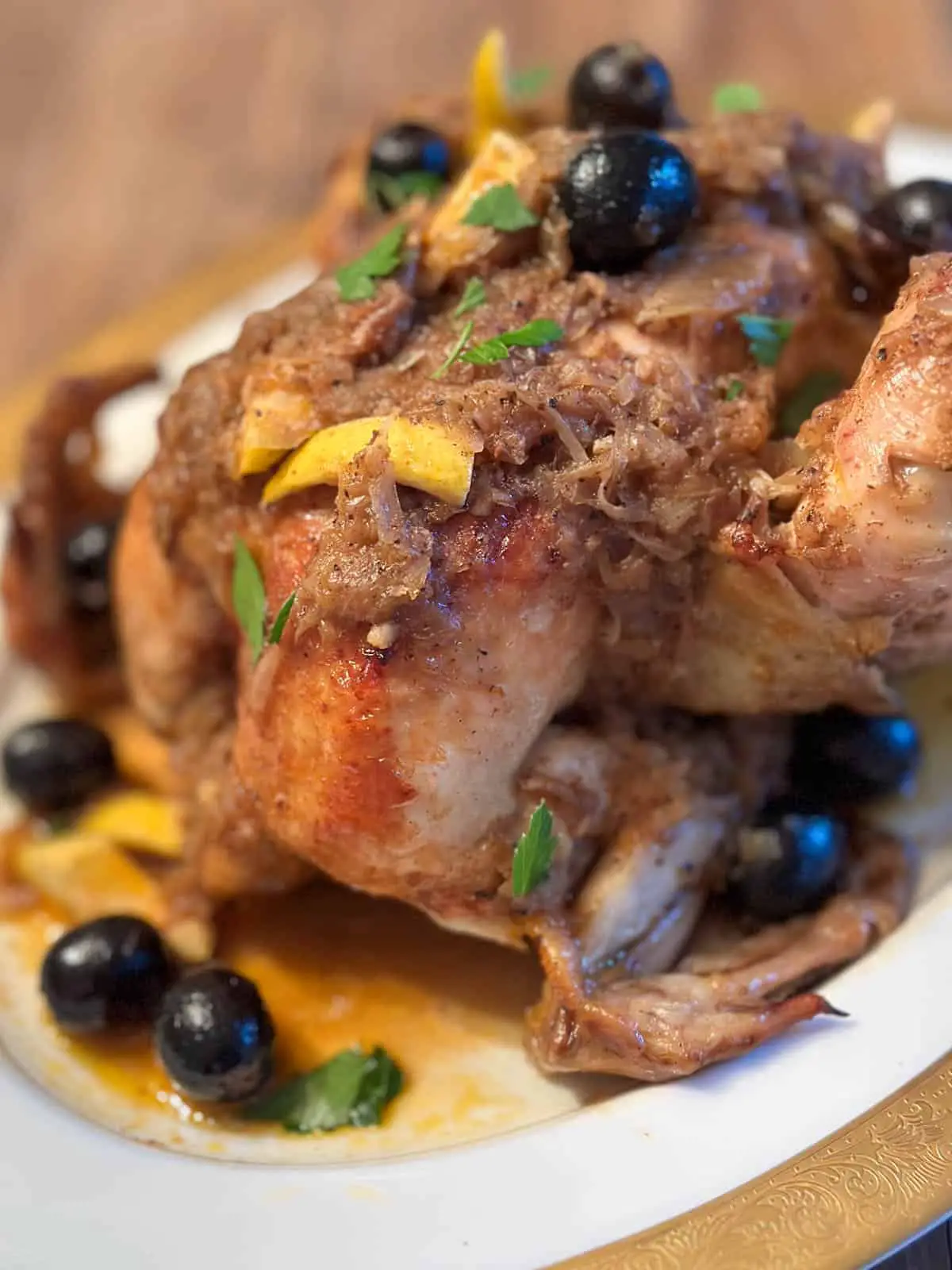 A whole chicken on a white platter rimmed with gold. The chicken is covered with an onion sauce, and there are olives, pieces of lemon rind, and Italian parsley as garnish.