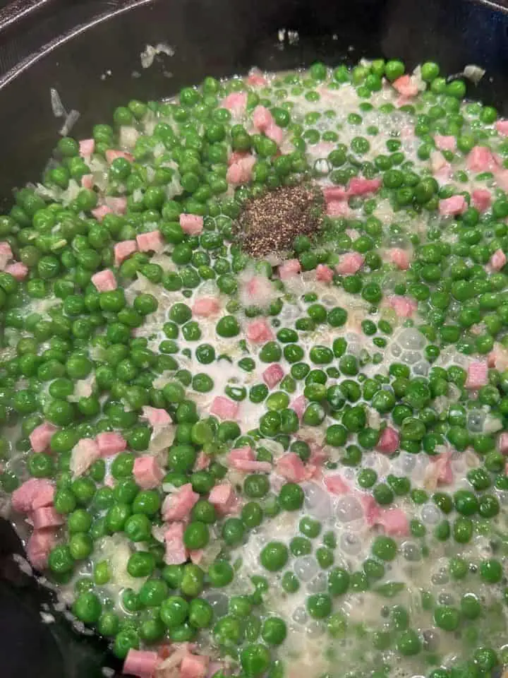Peas, diced ham, onion, and cream in a cast iron skillet. There is salt and pepper sprinkled on top.