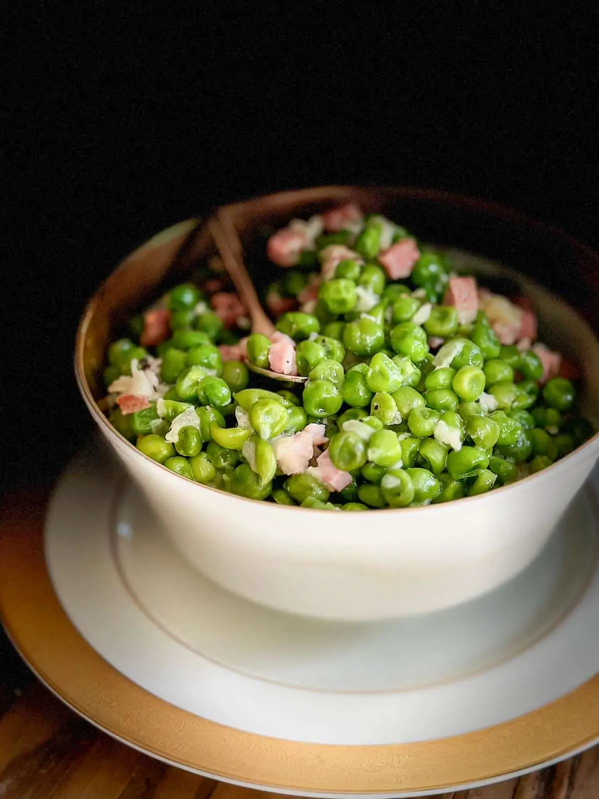 A gold rimmed bowl containing peas, ham, and onion with a gold stemmed spoon resting in the bowl. Part of the picture is in shadow giving it a moody look.