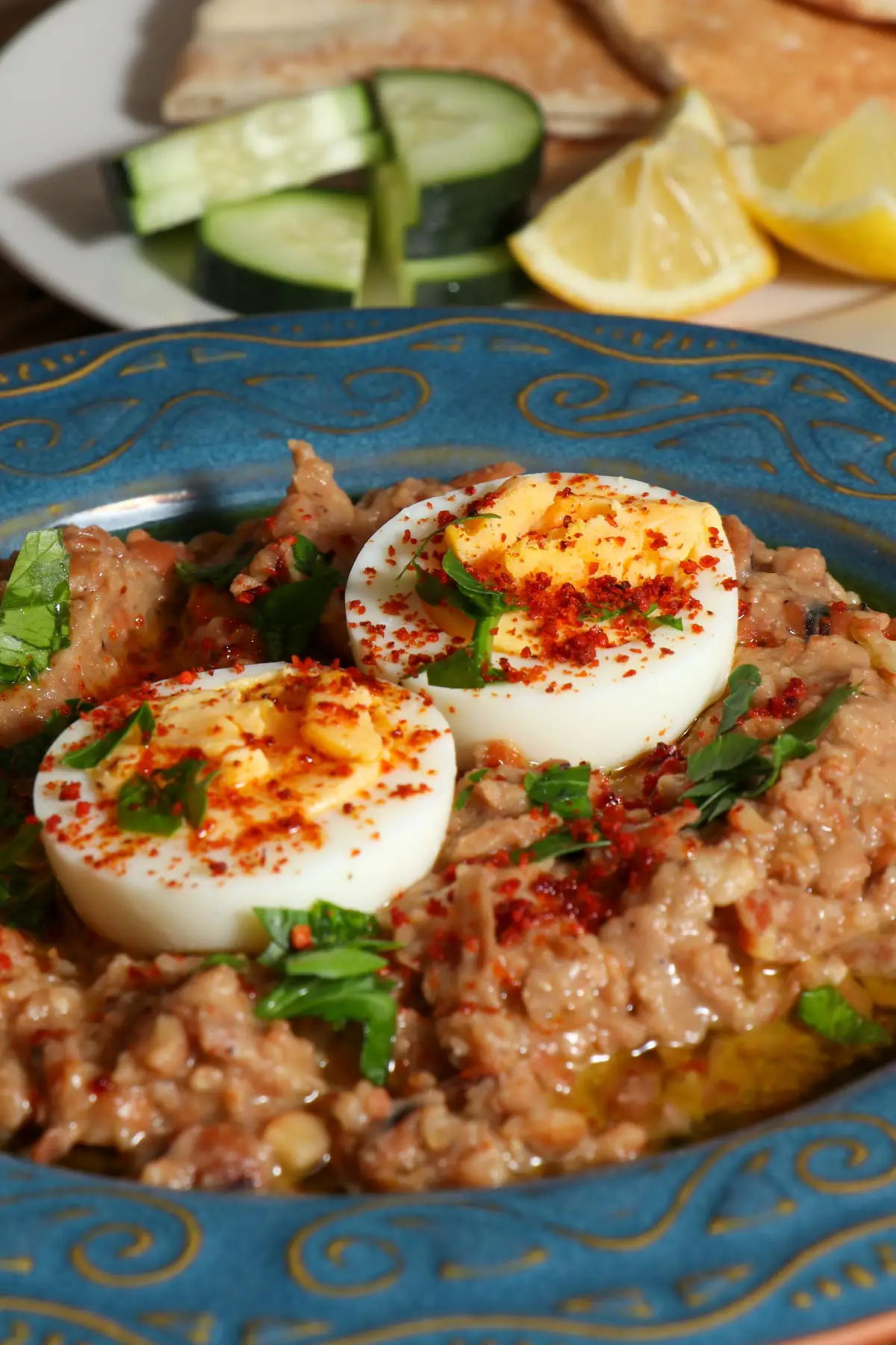 A blue patterned bowl containing foul which is made of mashed fava beans topped with olive oil, minced parsley, hard boiled eggs, and Aleppo pepper. There is a white plate in the background with sliced cucumber, pita bread, and lemon wedges.