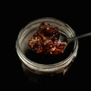 A jar that is mostly in shadow but you can see the top of the jar and above it is a spoon holding Thai chili oil and flakes.