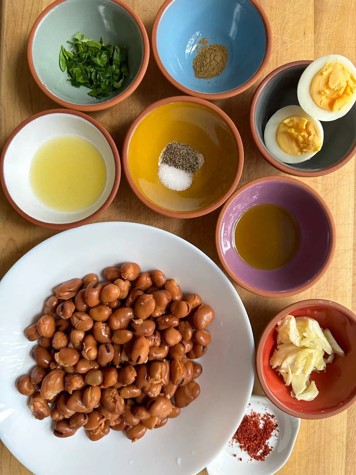 A white bowl with fava beans, small bowls with parsley, lemon juice, cumin, salt and pepper, hard boiled eggs, olive oil, and crushed garlic. There is a small white dish with red pepper flakes.