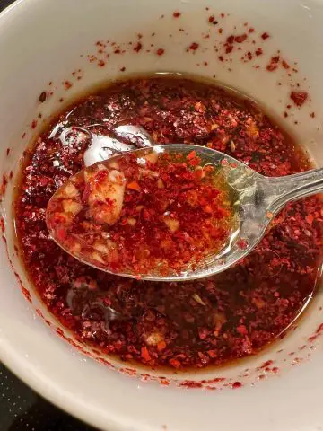 A white bowl containing spicy braising sauce including garlic and red pepper flakes. There is a spoon containing some of the sauce in the foreground.