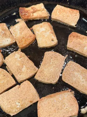 Rectangular pieces of tofu pan in a cast iron skillet being pan fried in oil.