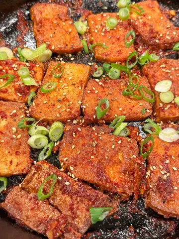 Pan fried pieces of tofu in a cast iron skillet that has been braised with a spicy sauce so that the pieces are red in color. The tofu is garnished with green onions and sesame seeds.