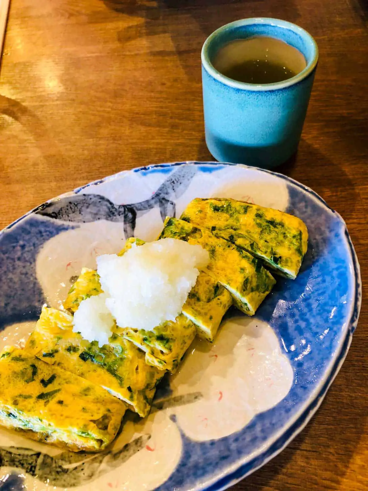 A Japanese omelette topped with whitebait on a blue and white plate with a blue coffee cup in the background. 