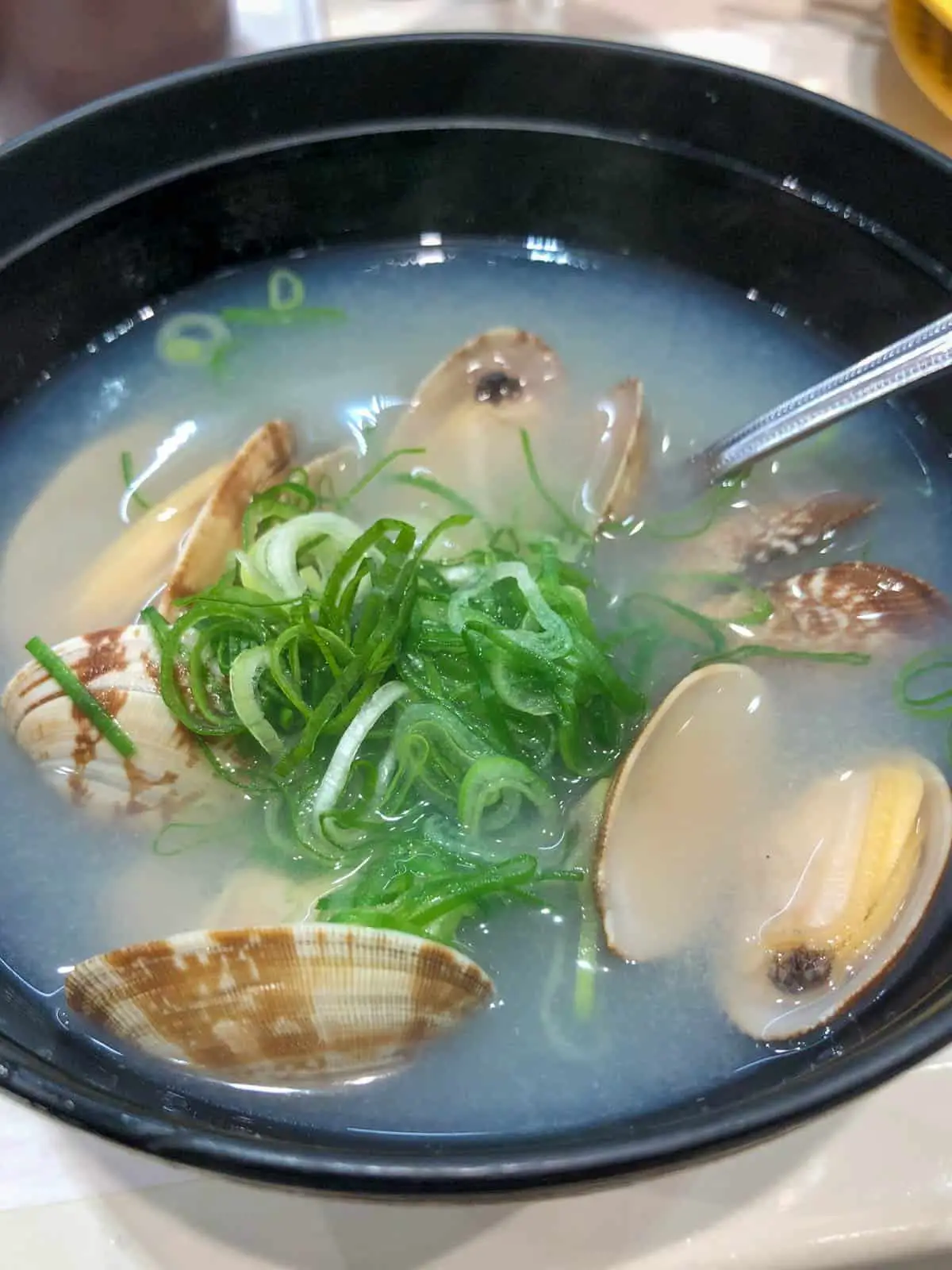 A black bowl containing miso soup with clams and garnished with green onions. There is a spoon resting in the bowl.