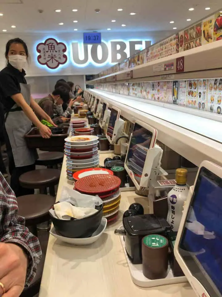 UOBEI restaurant in Tokyo with view of the counter where people are sitting and stacks of empty plates. There are screens where people order food and the conveyer belt above the screens. There is a lady with a mask in the picture.