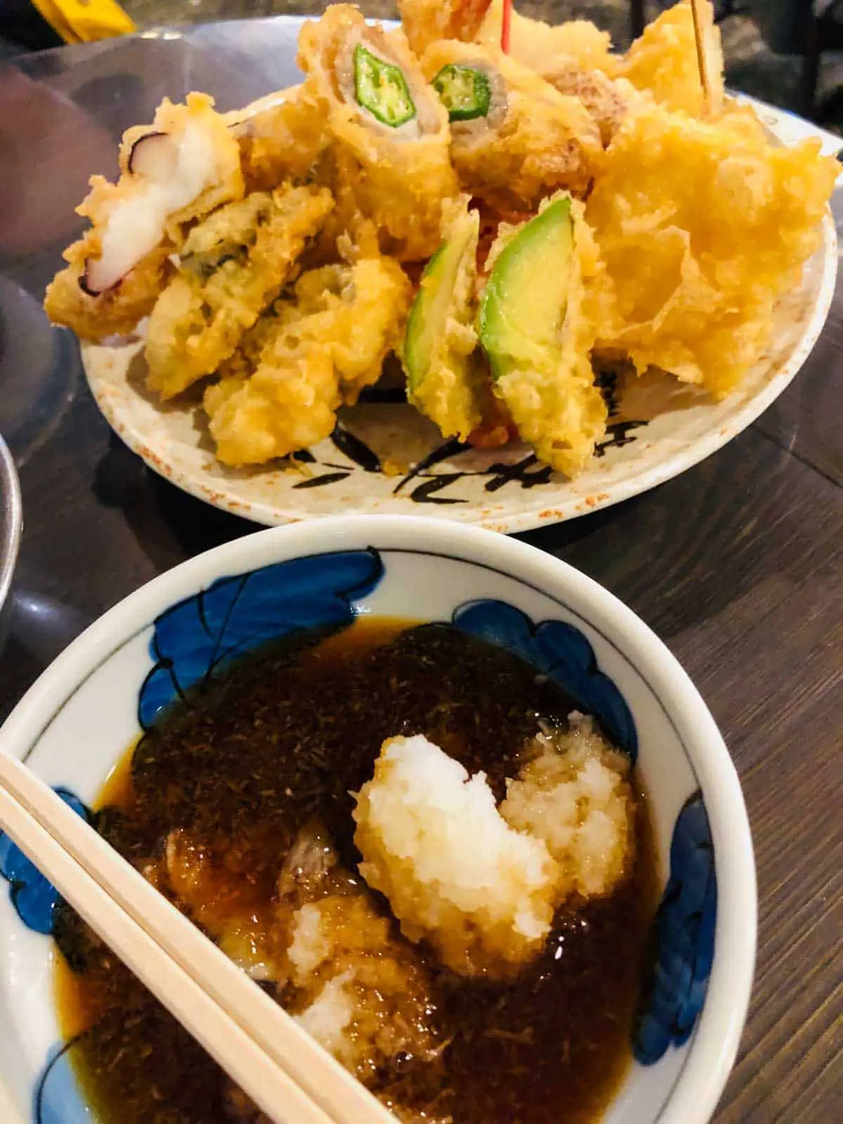 A dish topped with various pieces of tempura and a bowl with soy sauce and grated daikon radish with chopsticks resting on top.