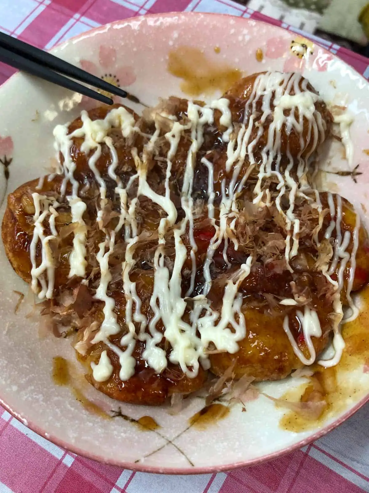 A plate full of takoyaki which is a ball shaped food filled with octopus and topped with drizzled mayonnaise and bonito flakes.  There is a pair of chopsticks resting on the side. 