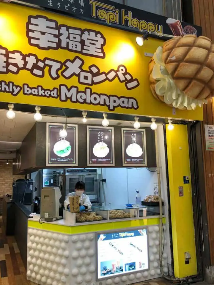 A Melon Pan Stand with yellow colored panels. There is a large fake melon pan bread on the upper panel of the stand and writing in Japanese as well as the words "Freshly Baked Melonpan" in English. There is a woman with a mask behind the stand.
