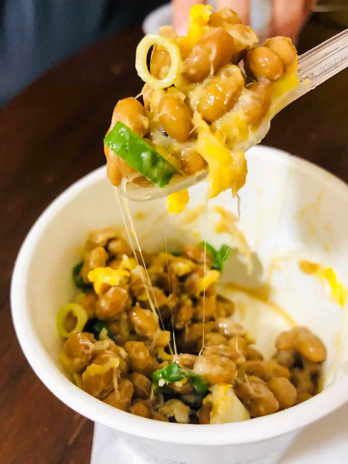 A cardboard cup with natto which is fermented soy beans.  Some natto is on a spoon and there is cooked egg and green onions, and slime. 
