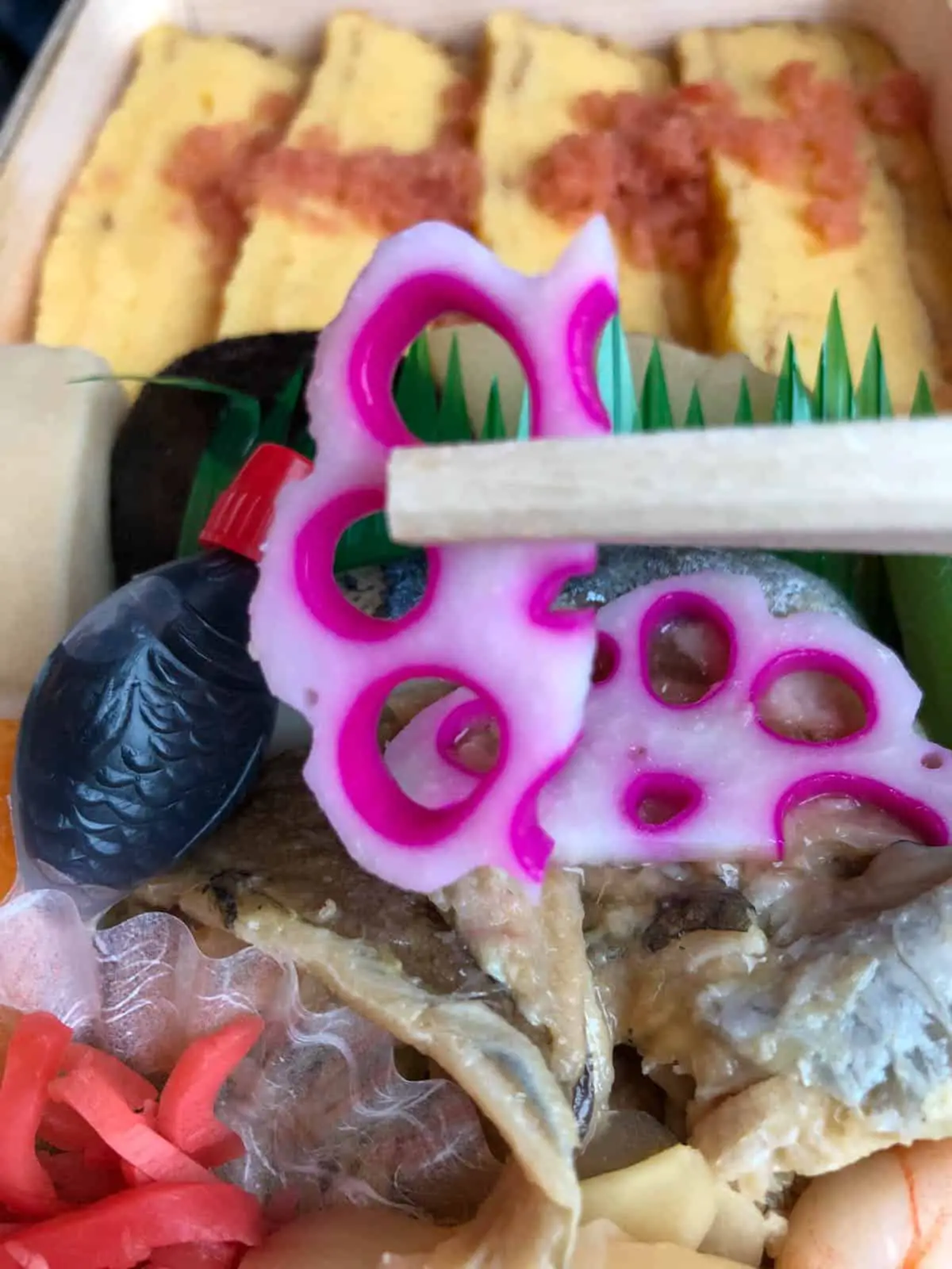 A chopstick holding a colorful pickled lotus root. There is also pieces of Japanese omelette, a fish shaped bottle of soy sauce, pickled ginger, and some fish in the bento as well. 