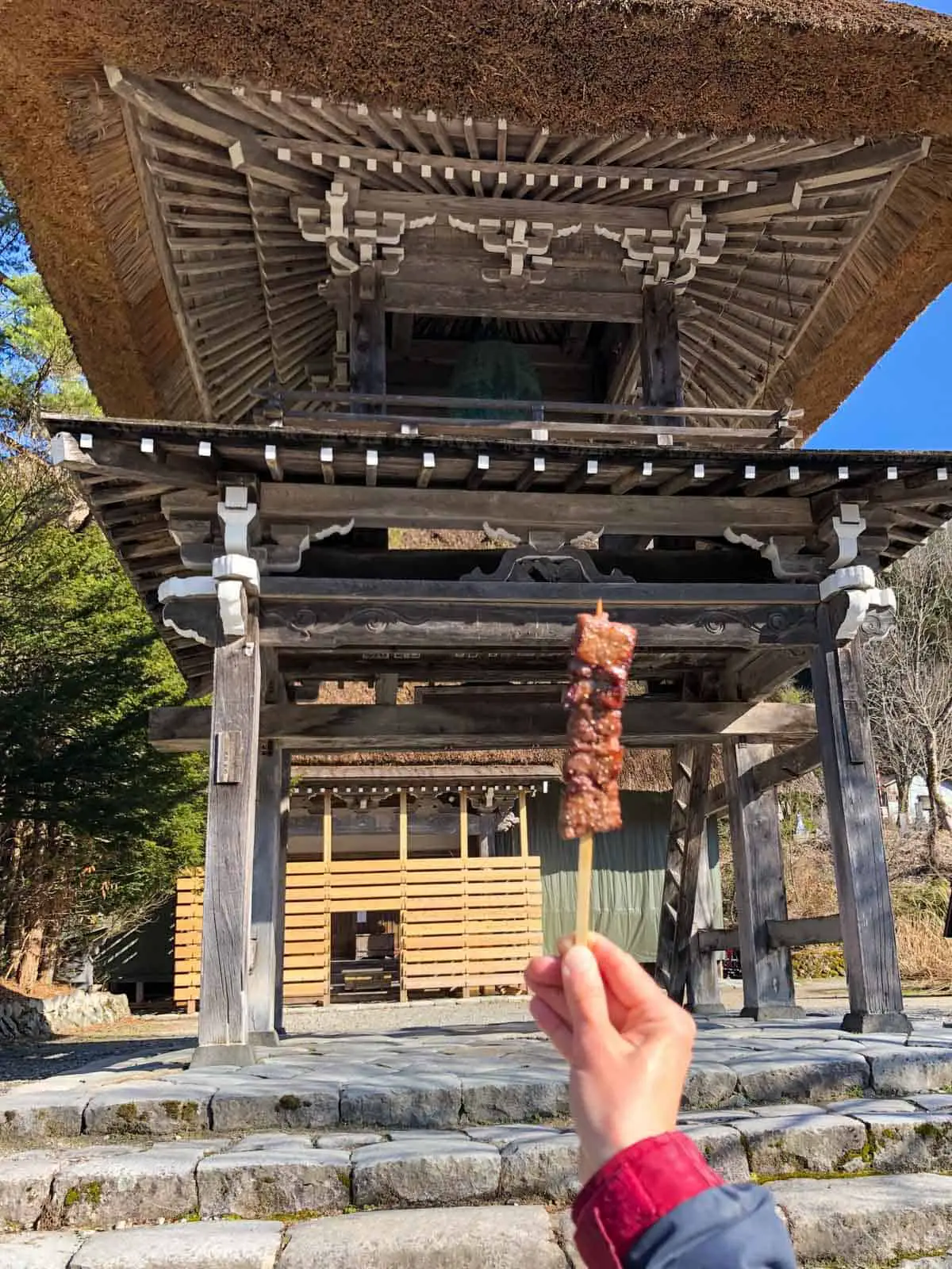 A temple in the background, and a hand holding a stick with beef skewered on it in the foreground. 
