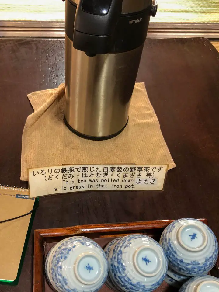 A thermos containing tea, some tea cups, and a sign that reads "This tea was boiled down wild grass in that iron pot" in English and Japanese.