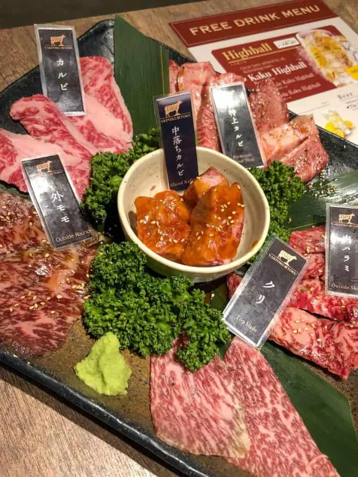 A selection of raw meats on a platter. Each meat has a sign with it and there are garnishes of parsley and a dab of wasabi on the plate.