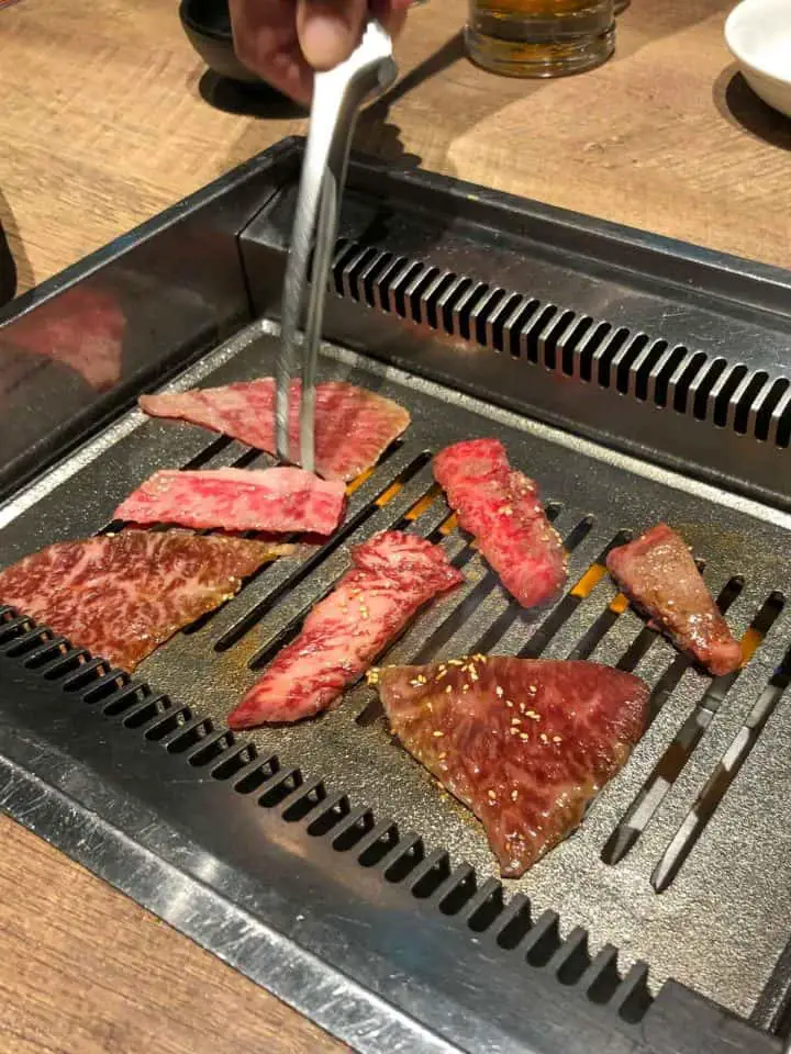A selection of sliced meats on an indoor table grill with a pair of tongs poised over one of the meats.