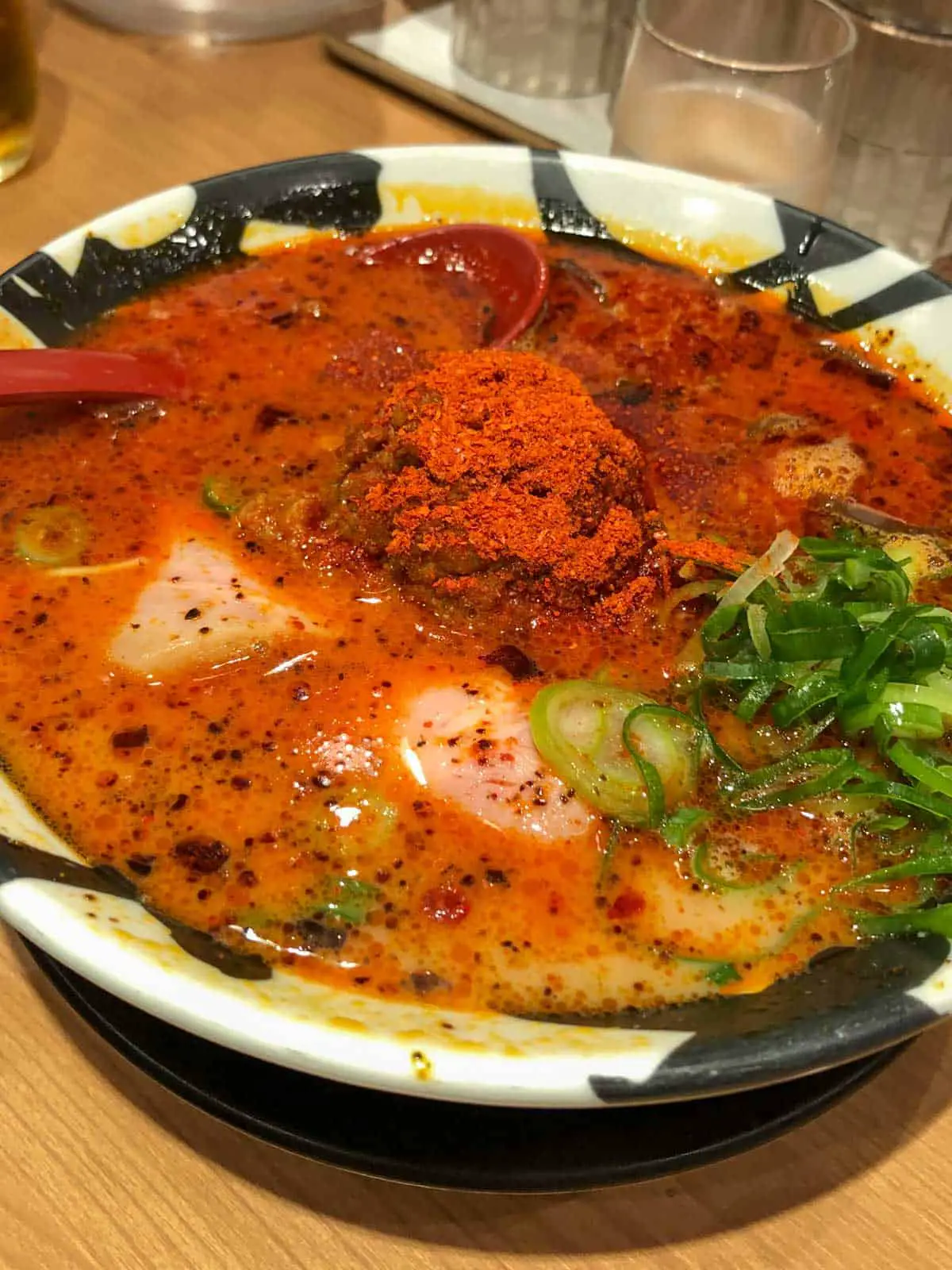 A bowl of super hot ramen with loads of red chili powder and sliced green onions. There is a red spoon resting in the bowl.