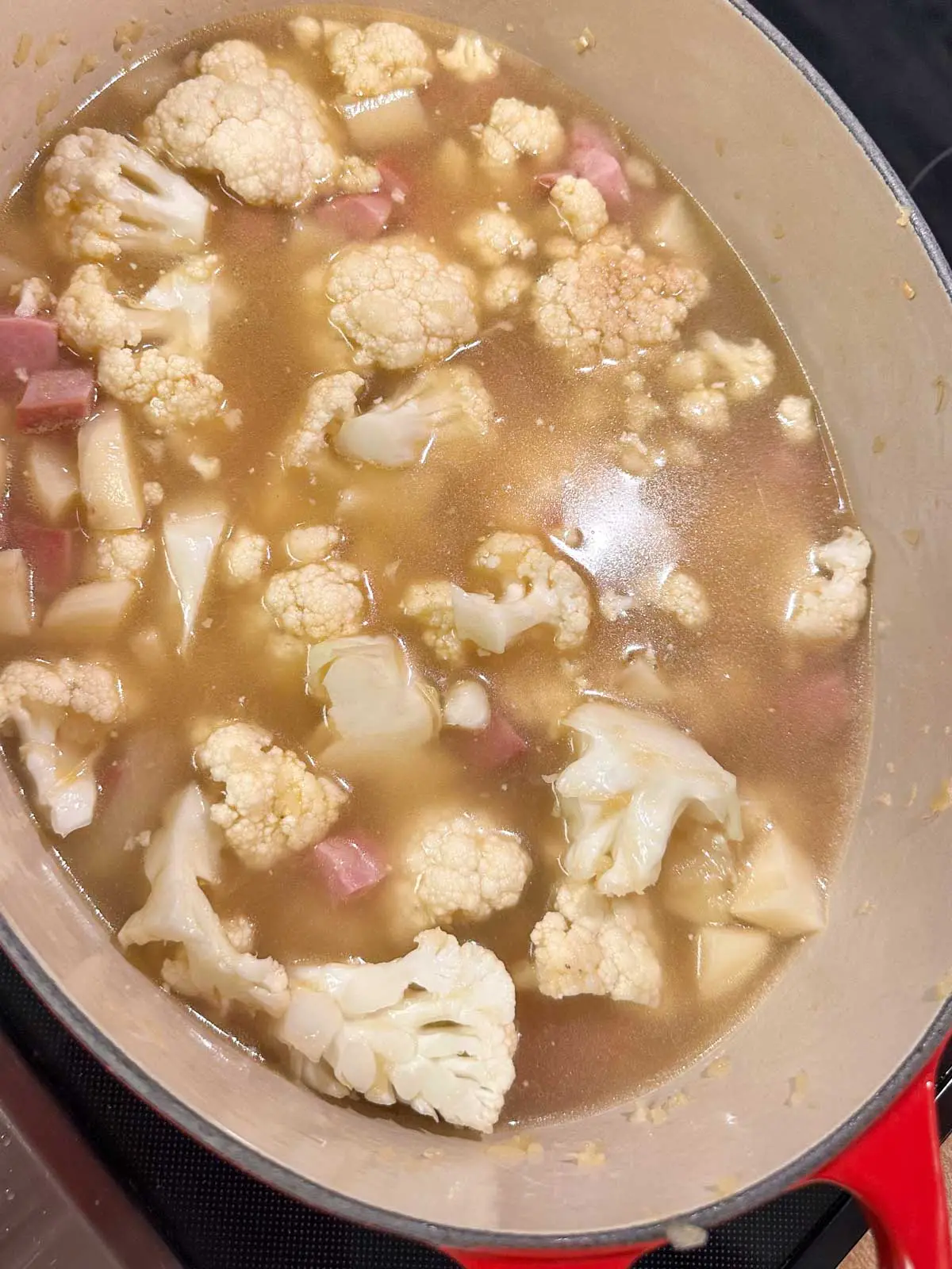 A red Dutch oven containing cauliflower florets, chicken broth, and chopped ham.