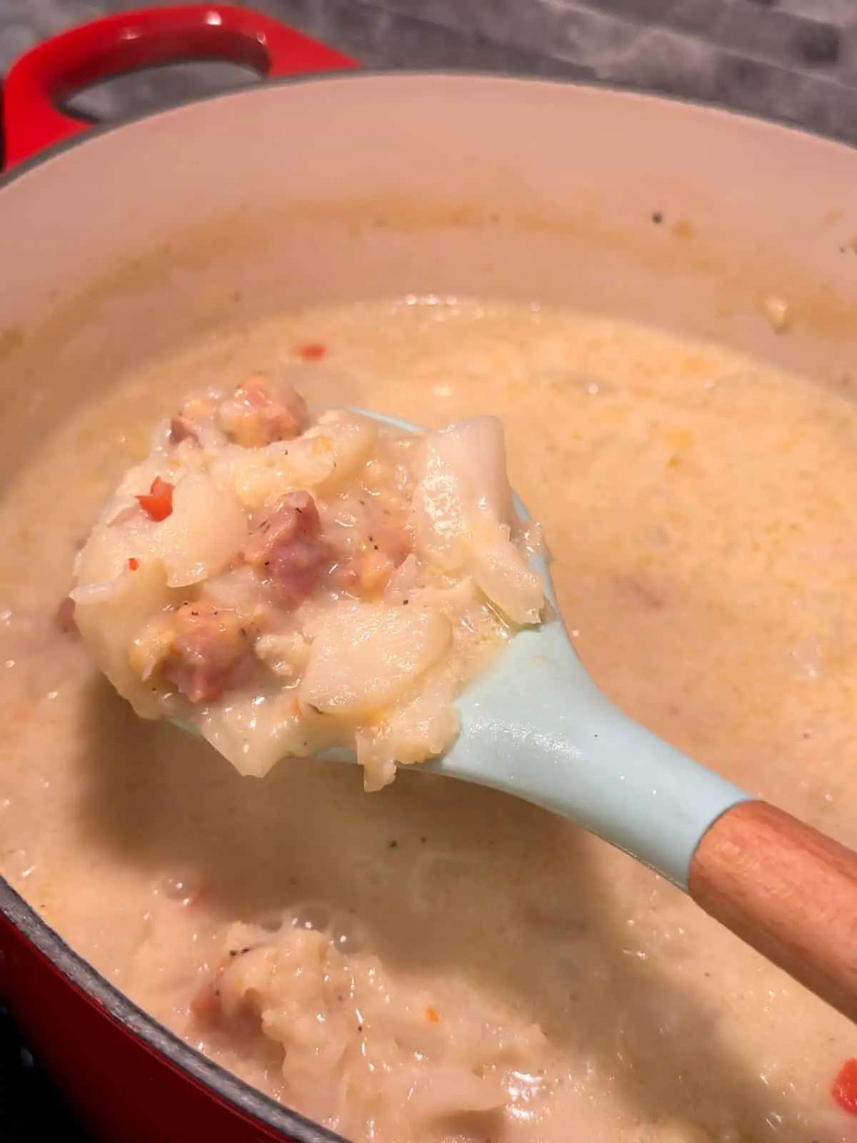 A red dutch oven containing a creamy cauliflower, potato, and ham soup. A blue silicone spoon with wooden handle contains some of the soup in the foreground.