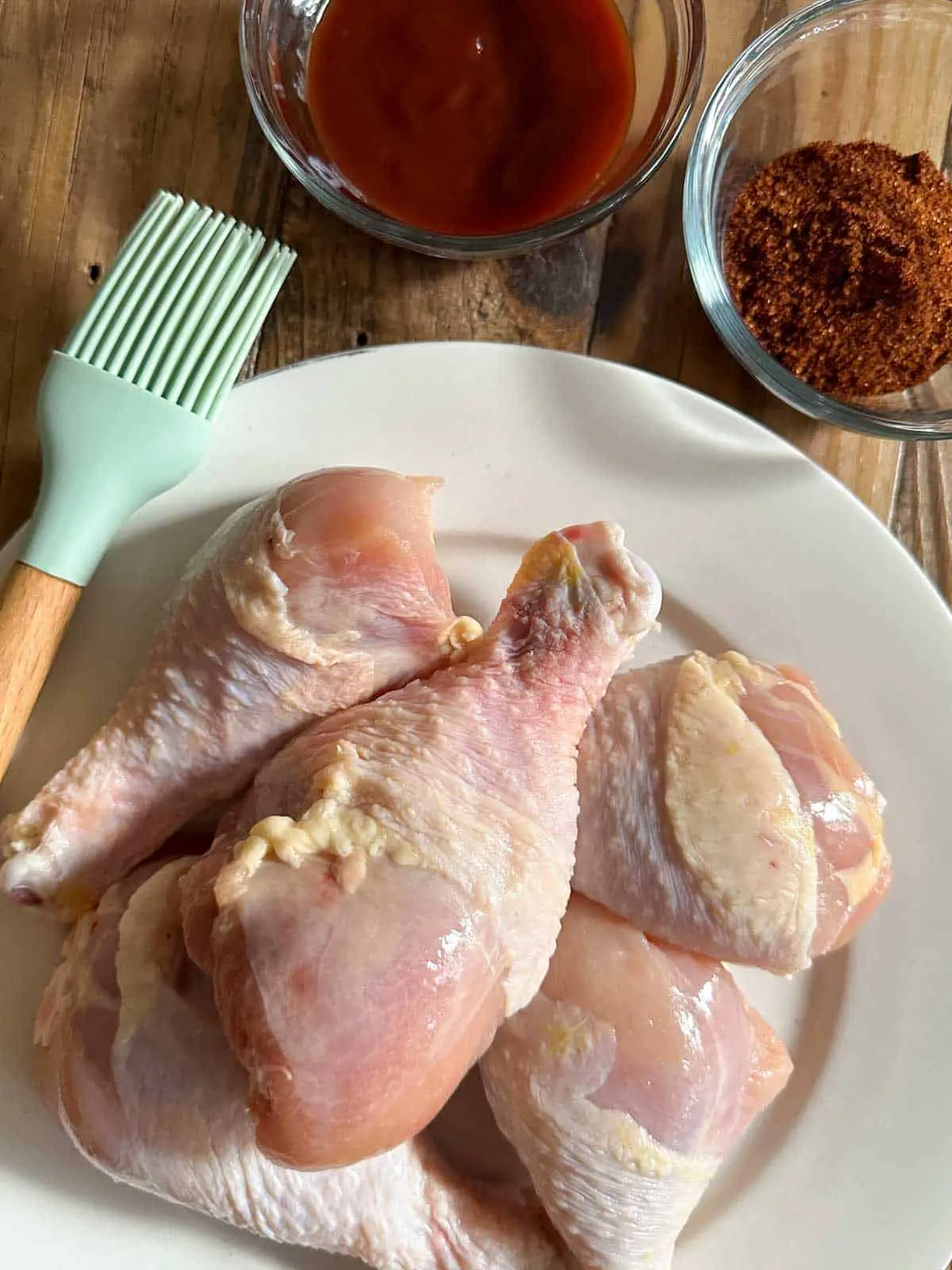 Raw chicken drumsticks on a white plate with a blue silicone utensil resting on the side of the plate, a bowl of barbecue sauce and a bowl of dry rub.