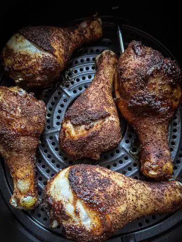 Seasoned chicken drumsticks which have been cooked in an air fryer in an air fryer basket. One of the drumsticks has a MEATER meat thermometer inserted.