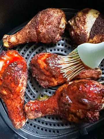 BBQ chicken drumsticks in an air fryer basket some are not basted with barbecue sauce and some have been basted with a blue silicone baster.