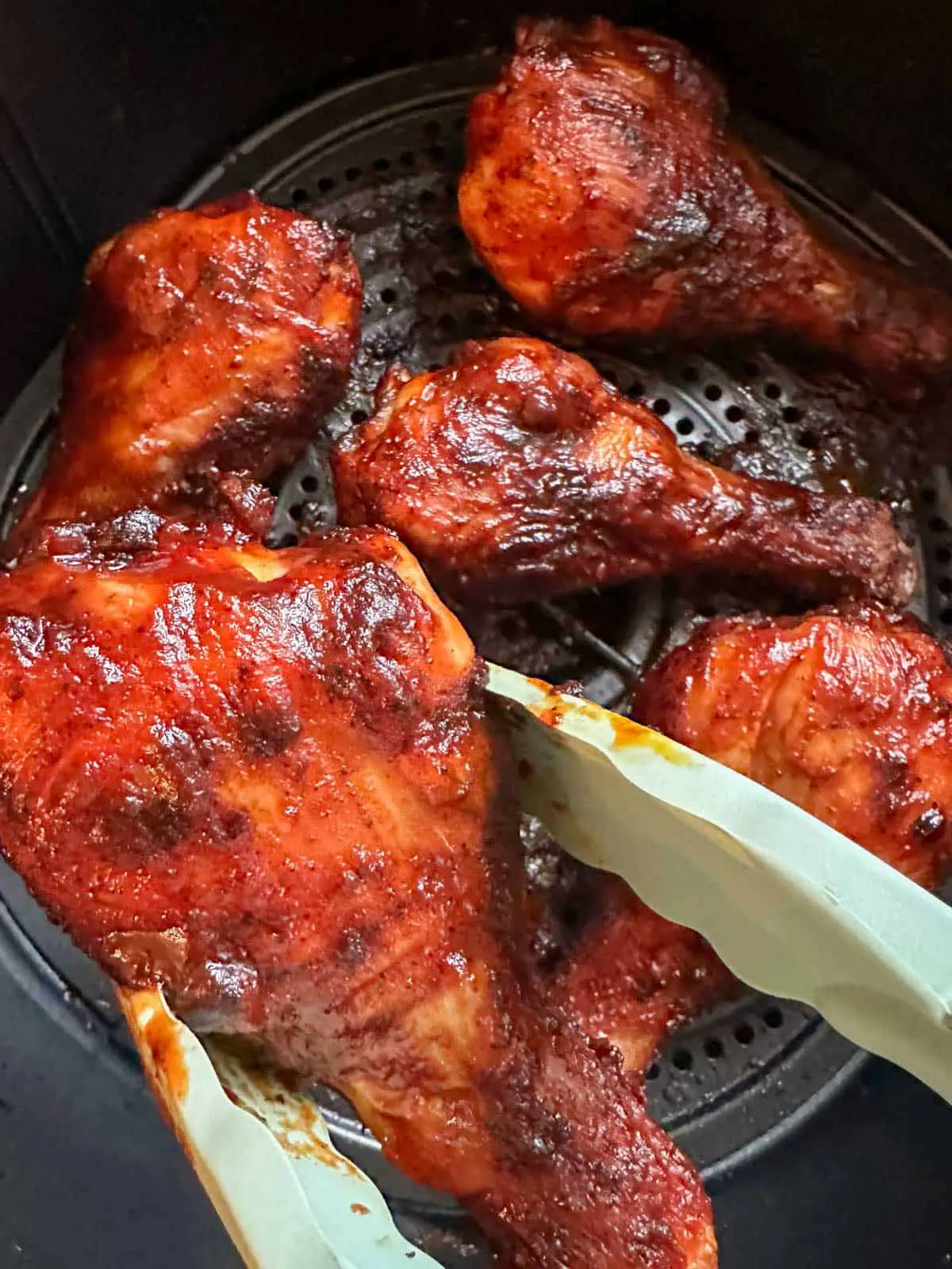 4 bbq chicken drumsticks in an air fryer basket with one being held by blue tongs in the foreground.