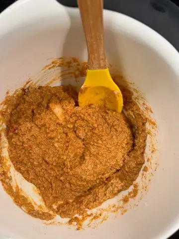 A white mixing bowl filled with a yellow colored yogurt based seasoning paste. There is a yellow spoon with a wooden handle resting in the bowl.