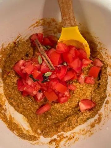 A white mixing bowl filled with a yellow colored yogurt based seasoning paste, diced tomatoes, a cinnamon stick, green cardamom, cloves, and black peppercorns. There is a yellow spoon with a wooden handle resting in the bowl.