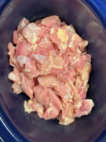 Bite sized pieces of raw chicken thighs in a blue slow cooker.