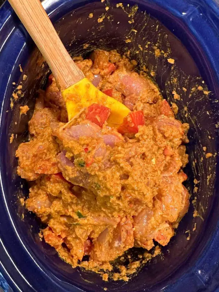 Raw chicken pieces coated with a yogurt based seasoning paste and tomatoes in a blue slow cooker with a yellow spoon with a wooden handle resting on the side.